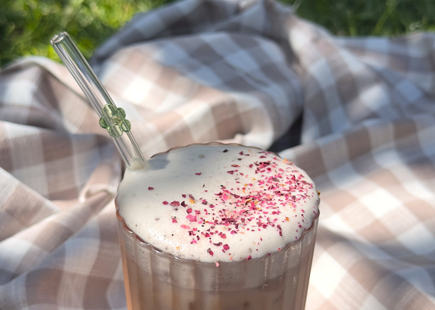 Brown gingham picnic cloth with a close up glass of foamy iced cherry chai. Crushed rose and cardamom dust the top.