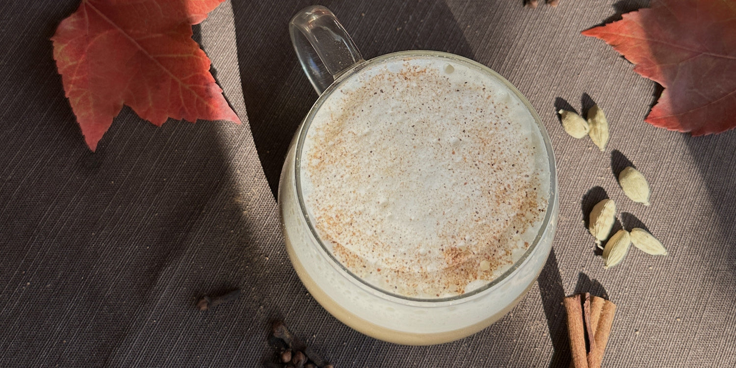 Birds eye shot of a clear mug with foamy pumpkin chai sitting on a brown tablecloth with soft sunlight and shadows. Red maple leaves, cardamom pods, and cinnamon sticks surround the mug.