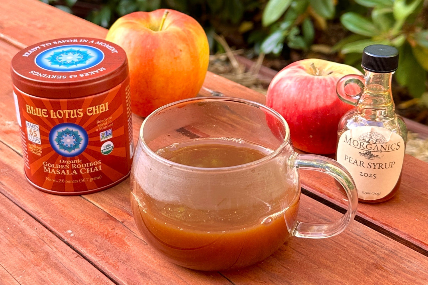 A clear mug of hot chai cider sits on a picnic table. Two apples are staggered behind it, with a small bottle of pear syrup to the right, and a tin of Golden Rooibos Masala Chai to the left.