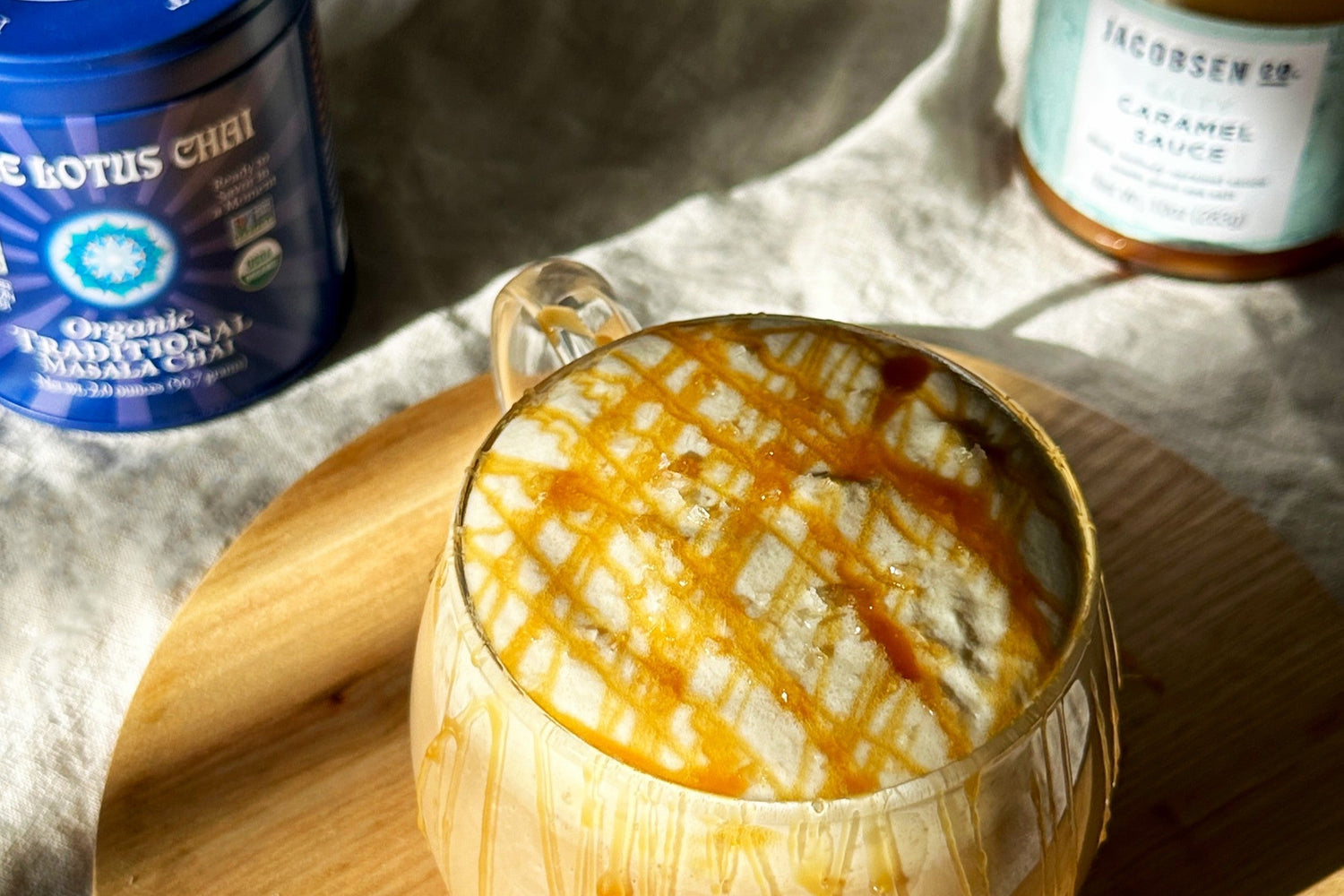 A caramel chai latte in a clear mug topped with cold foam and caramel drizzle. White background with a tin of organic traditional masala chai to the right and a jar of salty caramel sauce to the left.