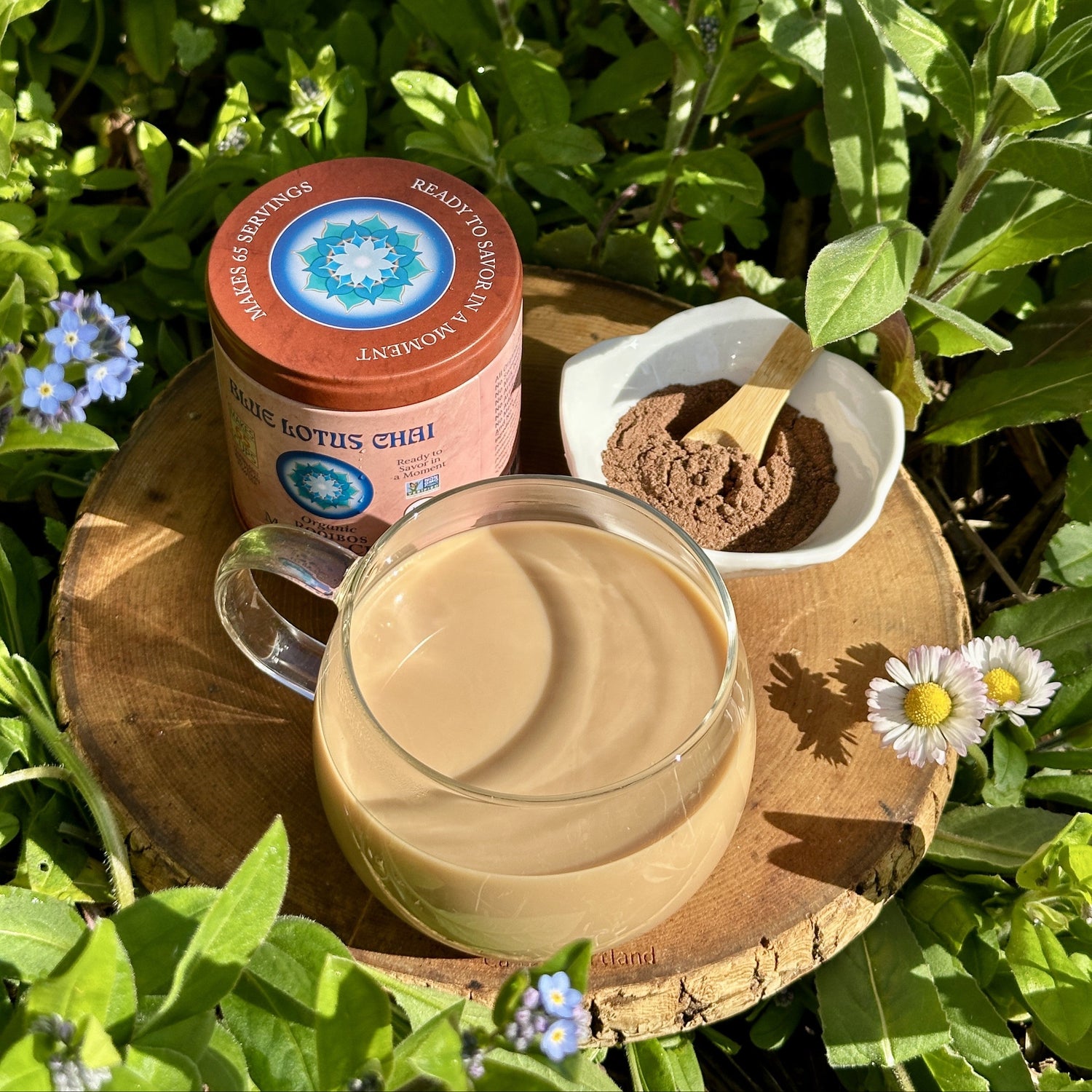 A glass mug filled with a warm chai sits on a round wooden tray outdoors among green leaves and small blue flowers. Next to it is a pink cylindrical container labeled ' Blue Lotus Chai' and a small white dish containing brown powder with a wooden spoon.