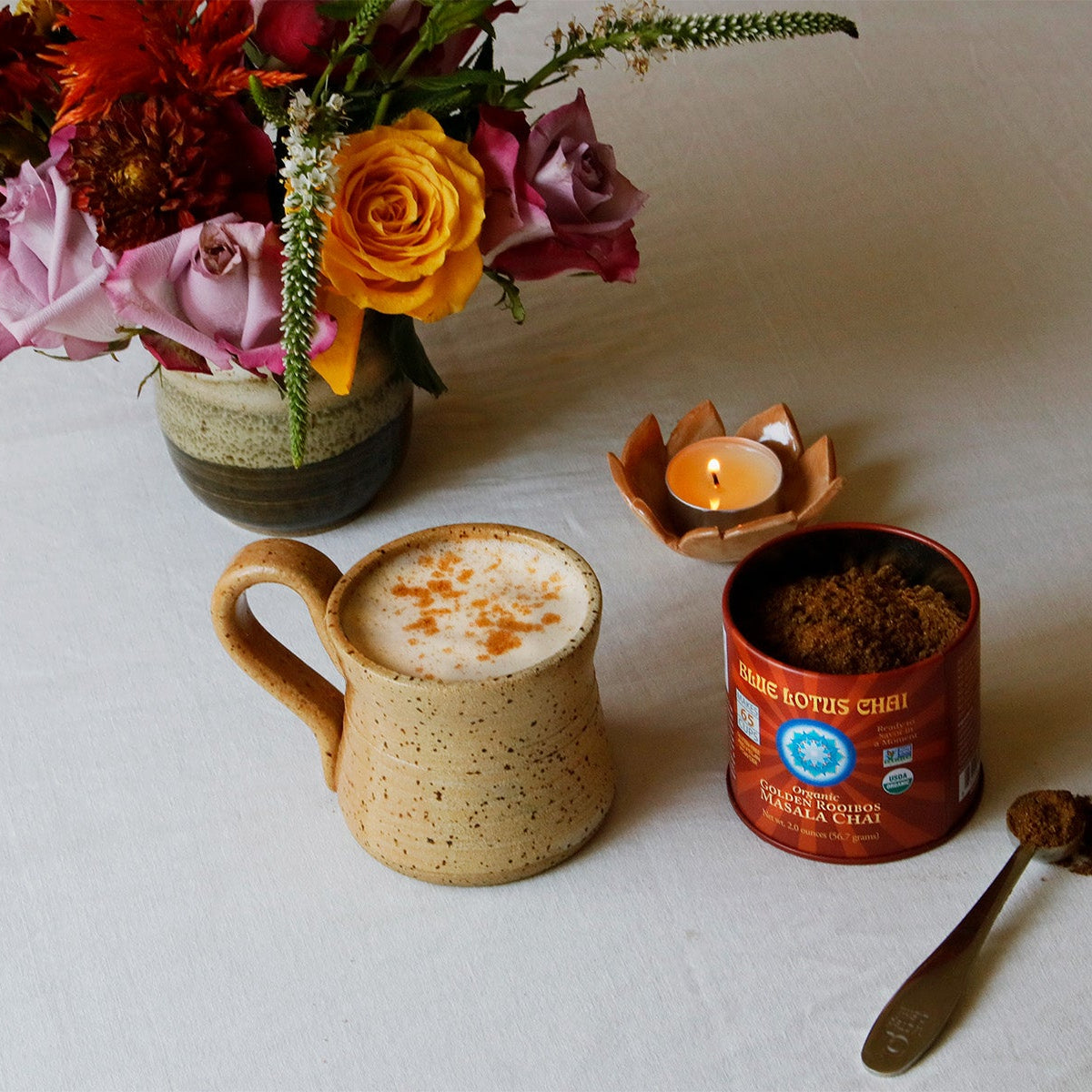 A styled tabletop scene on a white cloth featuring a speckled beige ceramic mug filled with a frothy hot drink topped with cinnamon, a red metal tin of coffee beans, a spoon with loose coffee, a small lit candle, and a flower arrangement in the background.