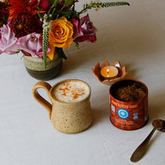 A styled tabletop scene on a white cloth featuring a speckled beige ceramic mug filled with a frothy hot drink topped with cinnamon, a red metal tin of coffee beans, a spoon with loose coffee, a small lit candle, and a flower arrangement in the background.