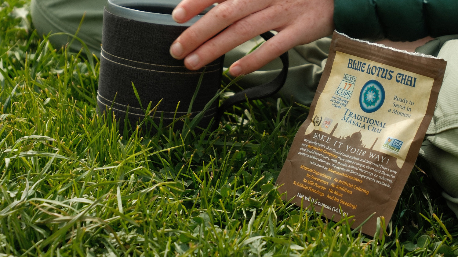 Person holding a black mug with a Blue Lotus Chai travel pouch on grass